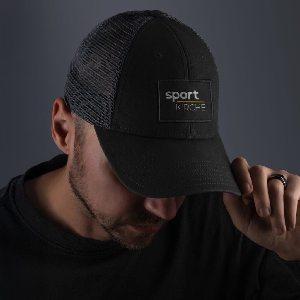 Person trägt schwarze Cap mit gesticktem „sportKIRCHE“-Logo auf der Vorderseite; neutraler Hintergrund.