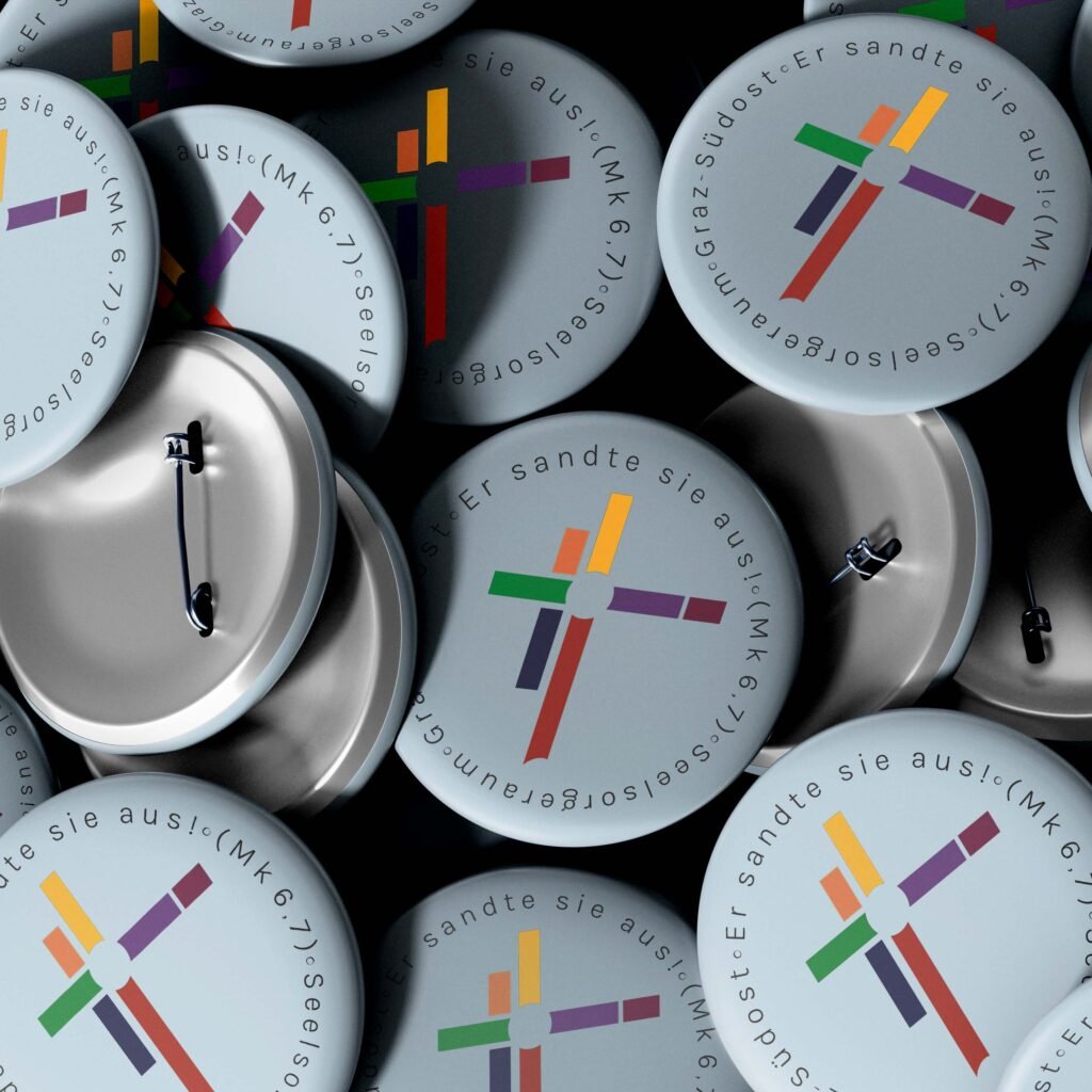 Mehrere runde Ansteck-Pins mit farbigem Kreuzlogo und dem Bibelzitat „Er sandte sie aus (Mk 6,7)“ – Seelsorgeraum Graz-Südost.