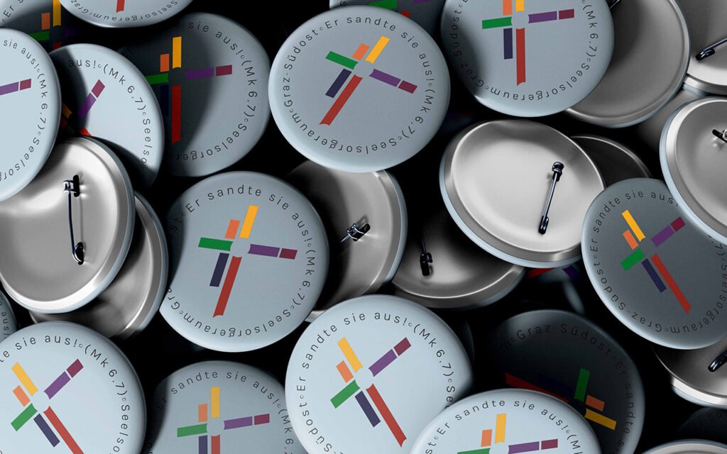Mehrere runde Anstecker mit Logo des Seelsorgeraums Graz-Südost – abstrahiertes Kreuz mit bunten Linien und Schriftzug
