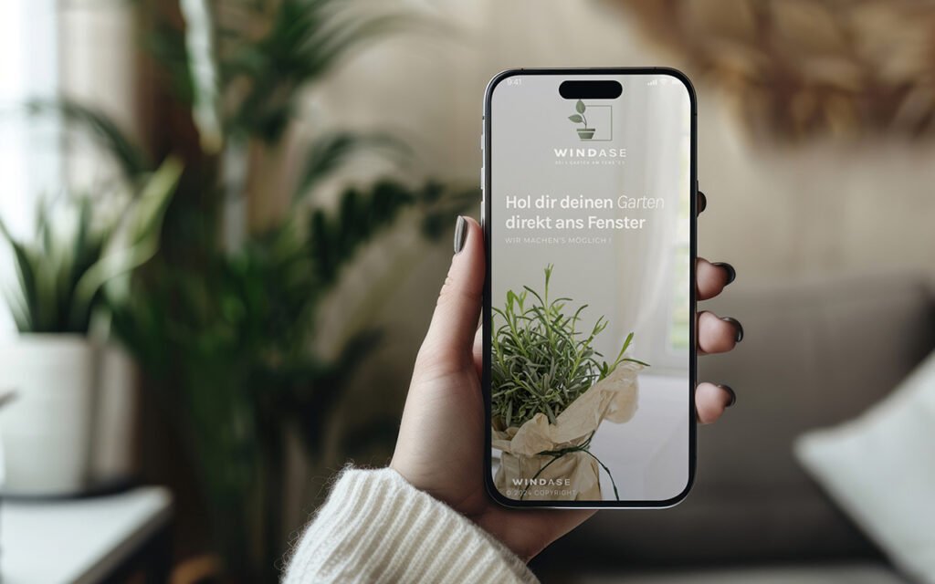 Smartphone in Hand zeigt Slogan „Hol dir deinen Garten direkt ans Fenster“ mit Pflanzen im Hintergrund – Branding-Ansicht für Urban Gardening-App Windase