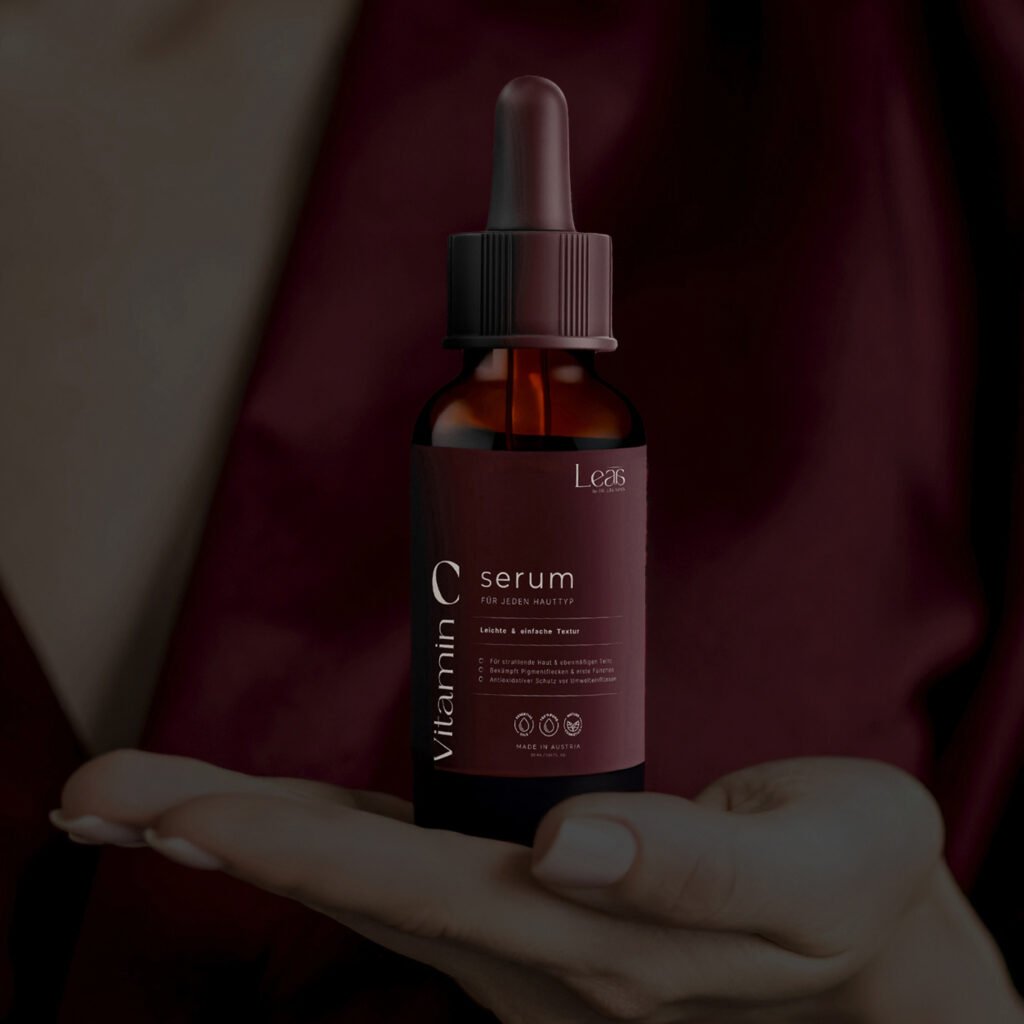 Hand präsentiert eine minimalistische Vitamin C Serumflasche vor einem bordeauxfarbenen Stoffhintergrund.