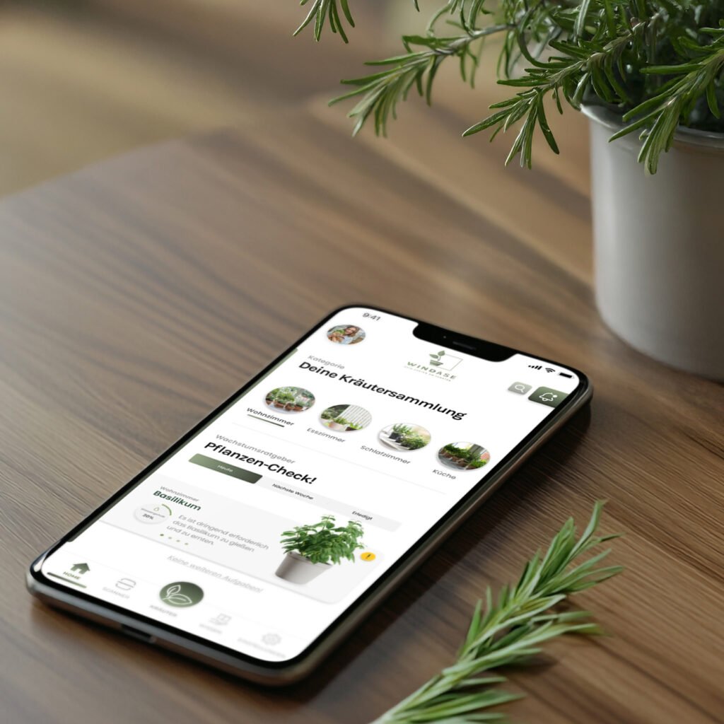 Smartphone mit geöffneter WINDASE-App auf einem Holztisch, im Vordergrund ein Rosmarinzweig und ein Kräutertopf.