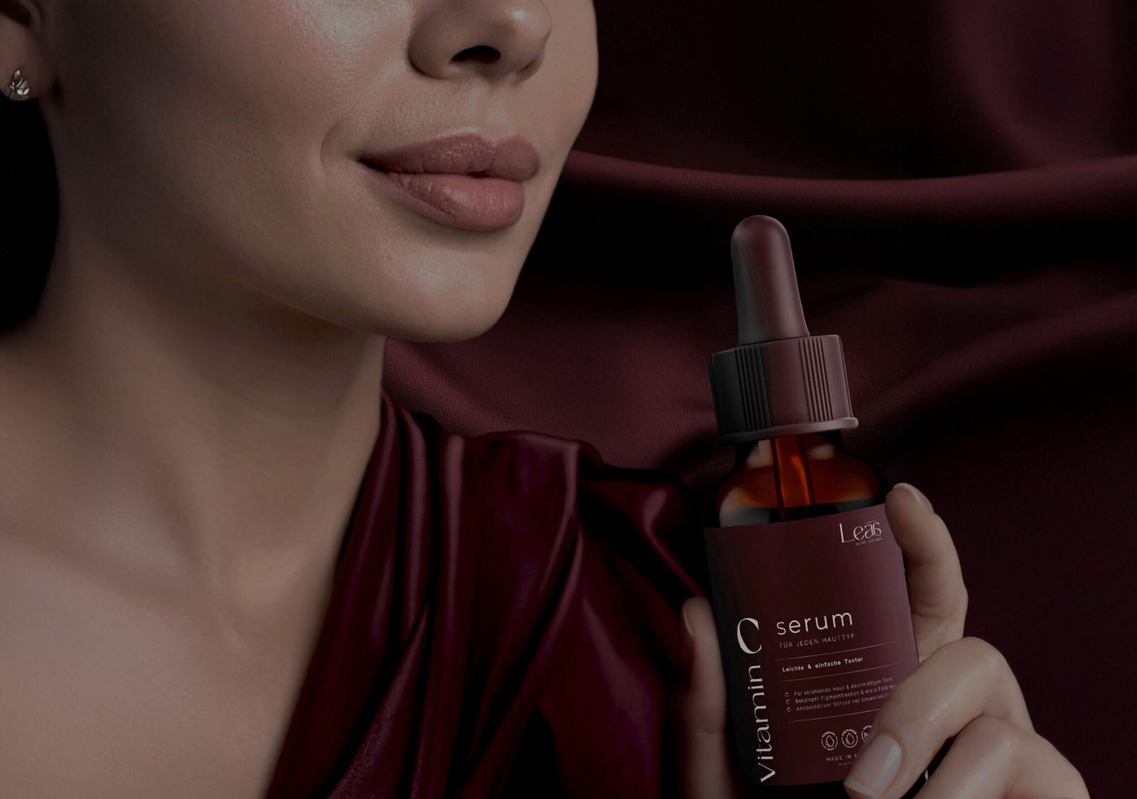 Nahaufnahme einer Frau mit glatter Haut, die ein Vitamin C Serum von Leas in einer dunkelroten Glasflasche hält. Hintergrund aus edlem, weinrotem Stoff. Beauty Produktfotografie – Logo Graz