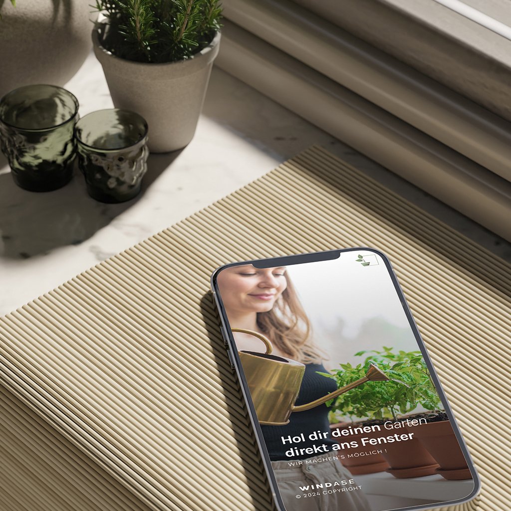 Smartphone mit geöffneter WINDASE-App auf einer hellen Oberfläche, im Bildschirm eine Frau, die Kräuter gießt.