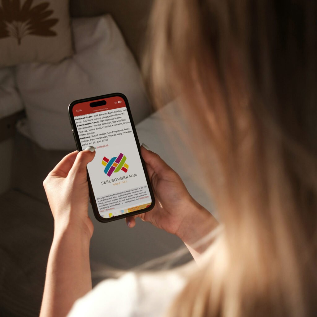 Frau hält Smartphone mit der Website des Seelsorgeraums Graz-Ost und farbigem Logo auf dem Display.