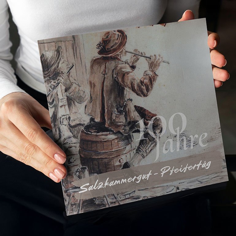 Person hält das Jubiläums-Booklet zum 100-jährigen Pfeifertag mit Illustration von zwei Musikanten.