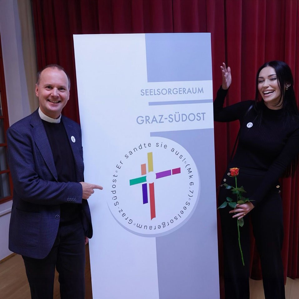 Zwei Personen präsentieren das neue Logo des Seelsorgeraums Graz-Südost auf einem großen Plakat – beide tragen passende Pins und lächeln in die Kamera. Linke Person: Stefan Ulz (Pfarrer), Rechte Person: Mateja Pejic (Designerin)