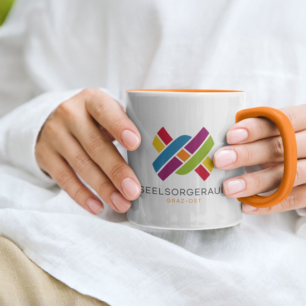 Weiße Keramiktasse mit orangefarbenem Henkel und dem bunten Logo des Seelsorgeraums Graz-Ost.
