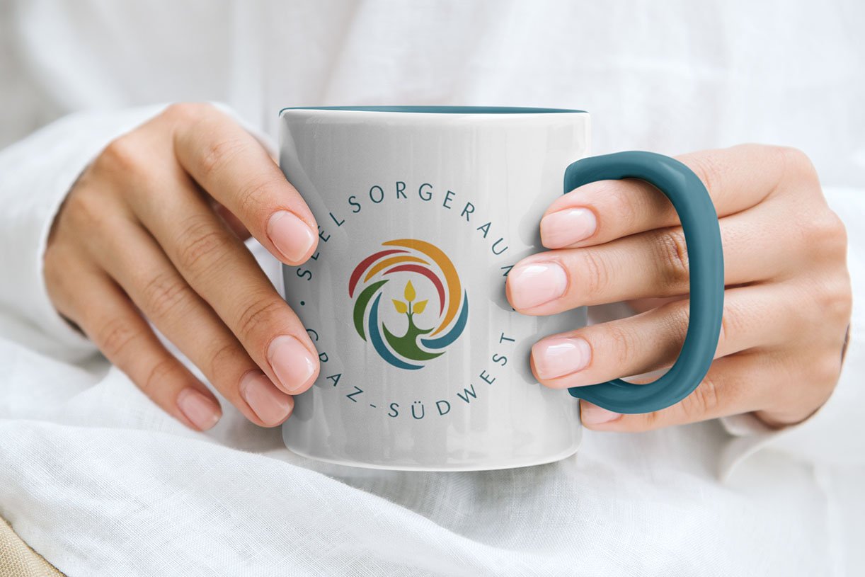Weiße Tasse mit türkisfarbenem Henkel und Logo des Seelsorgeraums Graz-Südwest, gehalten in beiden Händen.