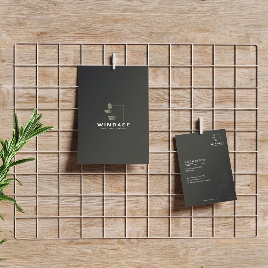 Poster und Visitenkarte mit WINDASE-Logo, platziert auf einem Metallgitter vor hellem Holz, dekoriert mit frischen Rosmarinzweigen.