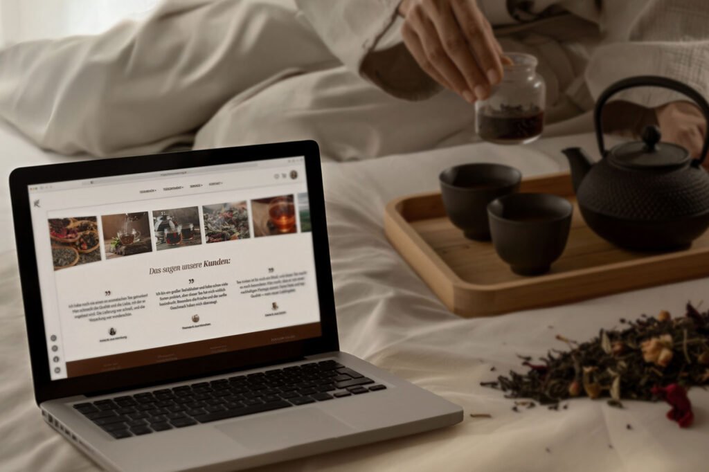 Laptop mit geöffneter Website einer Teemanufaktur, daneben Teekanne, Teetassen und Teeblätter auf einem Tablett – Darstellung von Webdesign im Alltag.