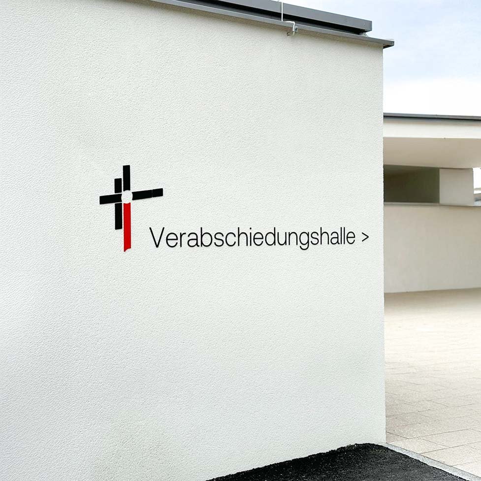 Wand mit Logo der Verabschiedungshalle am St. Peter Ortsfriedhof; schwarzes und rotes Kreuzsymbol mit Richtungspfeil.