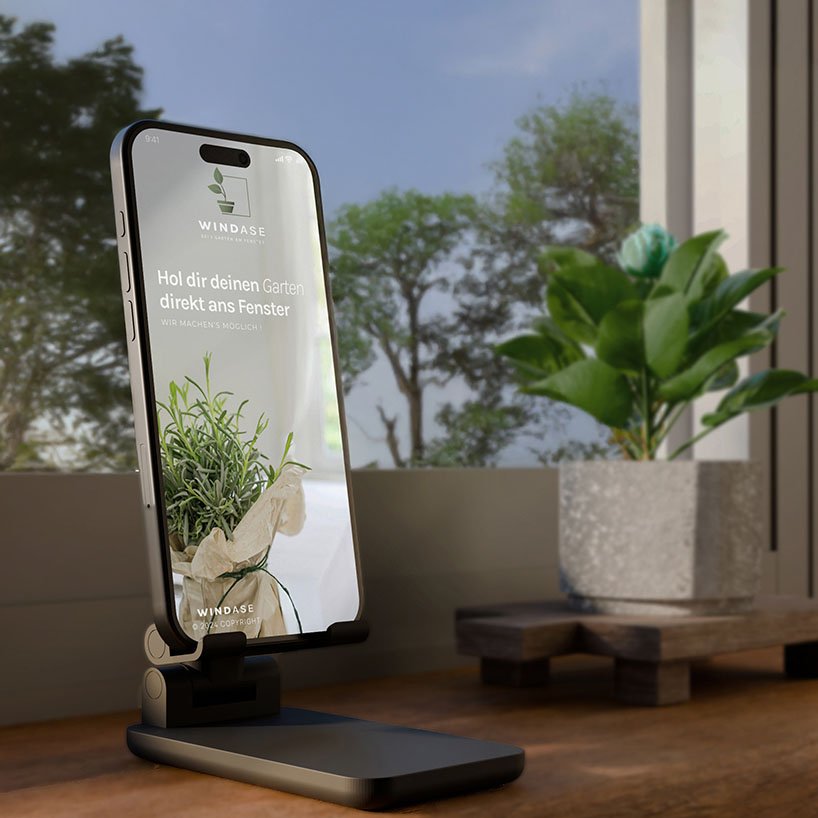 Smartphone mit geöffneter Windase-App, platziert vor Fenster mit Pflanzen – Indoor-Szenerie mit Urban Gardening Bezug