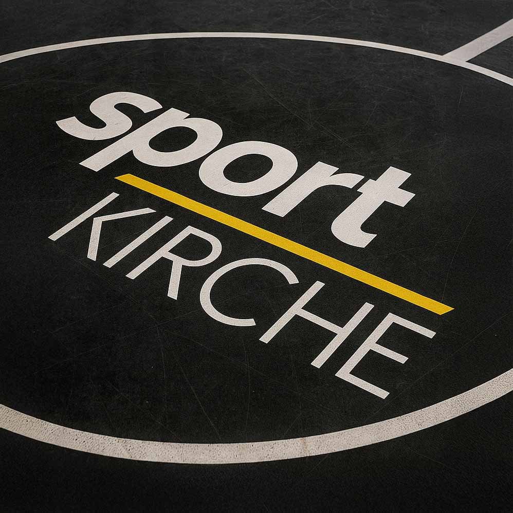 Logo „sportKIRCHE“ als Bodenmarkierung mit gelber Unterstreichung auf dunklem Sporthallenboden.